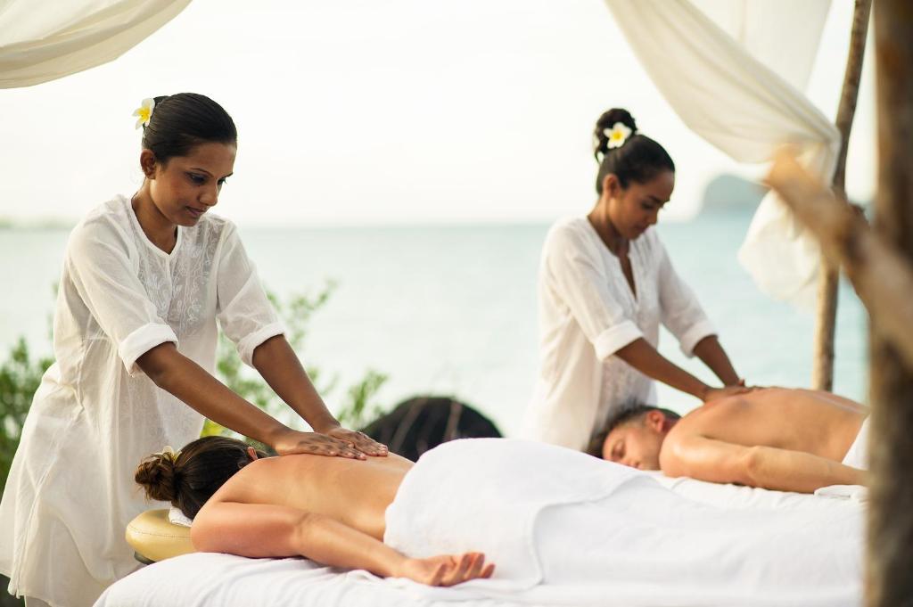 Divas masāžas terapeites nodarbojas ar masāžu pludmalē, sniedzot relaksāciju diviem klientiem. Fons ietver mierīgu jūras ainavu un dabu.