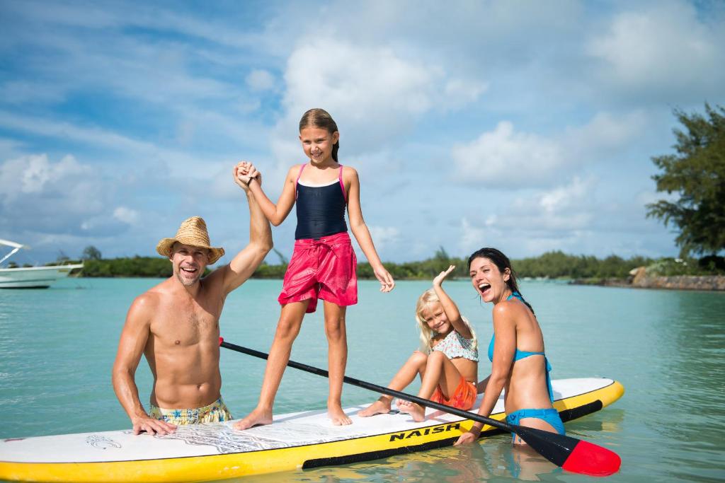 Ģimenes jautrība uz sērfinga dēļa ūdenī, kur divi bērni un pieaugušie smaidīgi izbauda sauli un brīvdienas.
