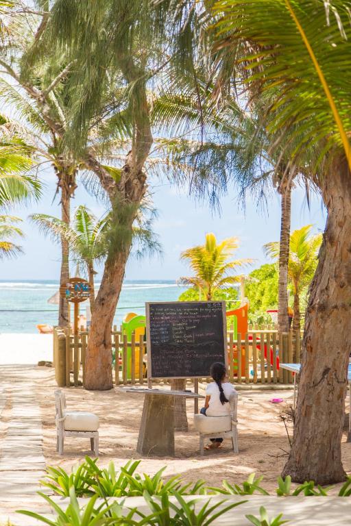 Pludmales ainava ar palmām, kur jauns cilvēks sēž pie galda ar melno tāfeli. Fons ietver jūru un bērnu spēļu laukumu.