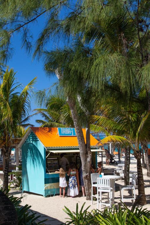 Kleita ar krāšņu pludmales kafejnīcu, kur bērni gaida pasūtījumu, ieskauta palmām un ziediem. Gaismas un krāsas rada priecīgu atmosfēru.