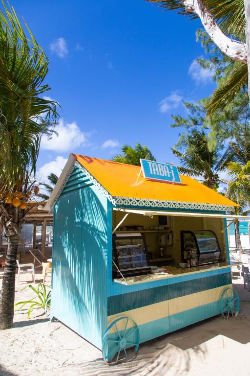 Pludmales kafejnīca ar gaišu zilu un oranžu jumtu, ko ieskauj palmas un skaidrs debesis. Kafejnīcā ir redzami galdi un saldējuma vitrīnas.