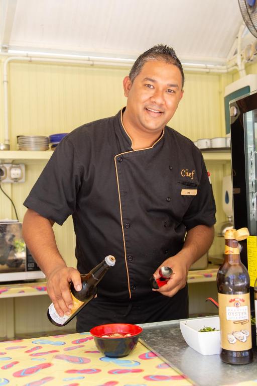 Šefpavārs gatavo ēdienu virtuvē, turēdams pudeles rokās un ar smaidu. Uz galda ir sarkana bļoda un citas sastāvdaļas.