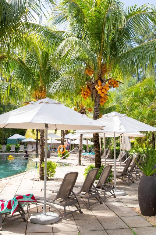Peldbaseins ar palmām un saulessargiem, kas nodrošina ēnu, radot relaksējošu atmosfēru. Apkārt redzami atpūtas krēsli un tropiskā vide.