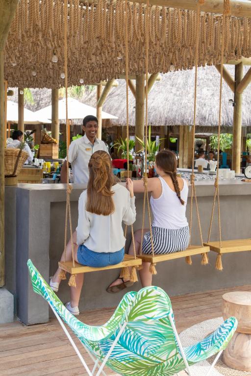 Divas sievietes sēž uz šūpuļtīkla, sarunājoties ar viesmīli, kas stāv pie bāra letes tropiskā vidē. Apsārtusi mājas interjers un tropu augi rada relaksējošu atmosfēru.