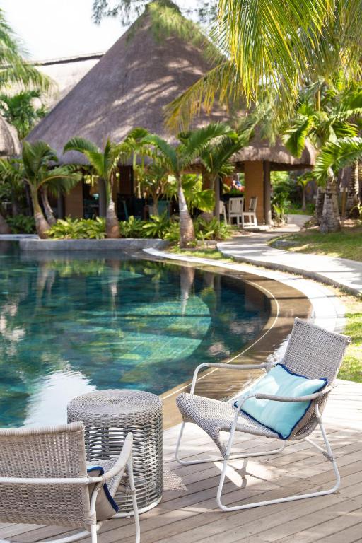 Mierīga atpūtas zona pie peldbaseina ar tropisko ainavu, kurā redzami vītņoti krēsli un galdiņš, kas aicina uz relaksāciju. Peldbaseina ūdens atspoguļo apkārtējās palmas un mājīgu mājiņu.