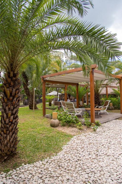 Zaļš dārzs ar palmām un ērtām atpūtas mēbelēm zem pergolas, kas piedāvā mierīgu atmosfēru. Akmens celiņš ved uz atpūtas zonu.