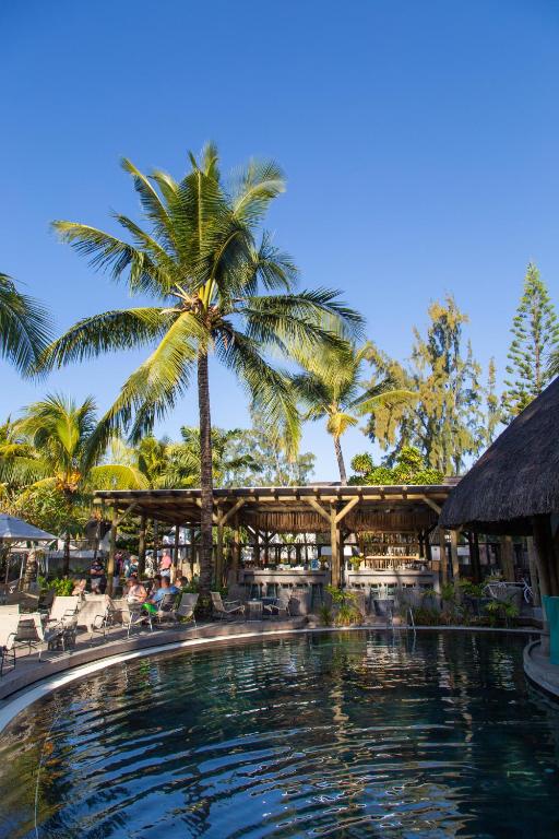 Tropu ainava ar palmām un baseinu, kur apmeklētāji bauda sauli un atpūtu. Fonā redzams bārs ar jumtu un zaļiem augiem.