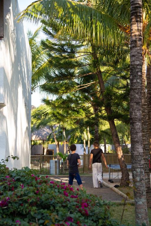 Divi bērni pastaigājas pa dārzu, apkārt augstiem palmu kokiem un krāšņām puķēm. Māja redzama attālumā, radot mierīgu un dabisku atmosfēru.