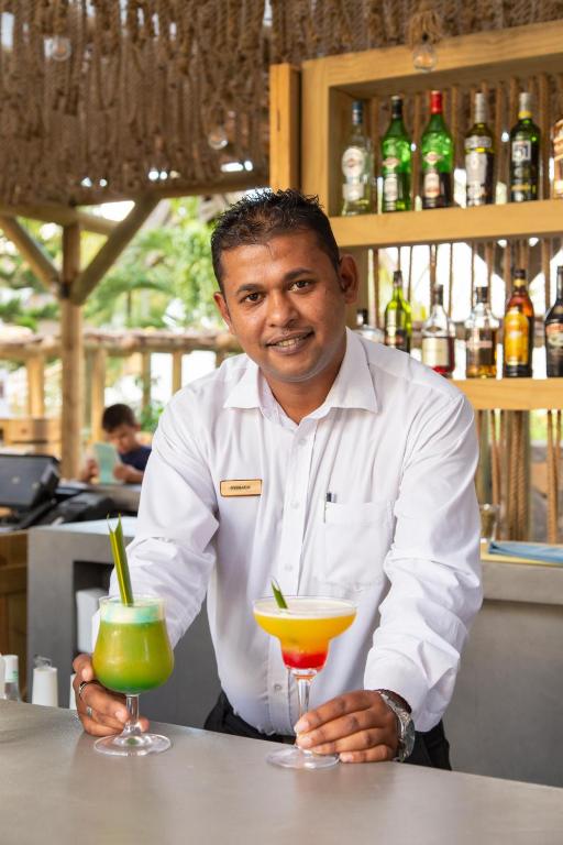Bārmenis, turot divus krāsainus kokteiļus, pozē pie bāra. Fons ietver dzērienu plauktu ar dažādām alkoholiskajām pudelēm.