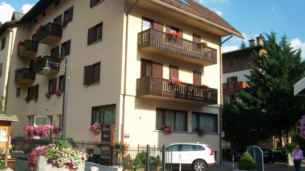 Trīs stāvu viesnīca ar balkoniem, kuriem piekārtas ziedošas puķes. Priekšā redzama auto stāvvieta un apzaļumotas teritorijas. Viesnīcas nosaukums ir "Garnì San Lorenzo".