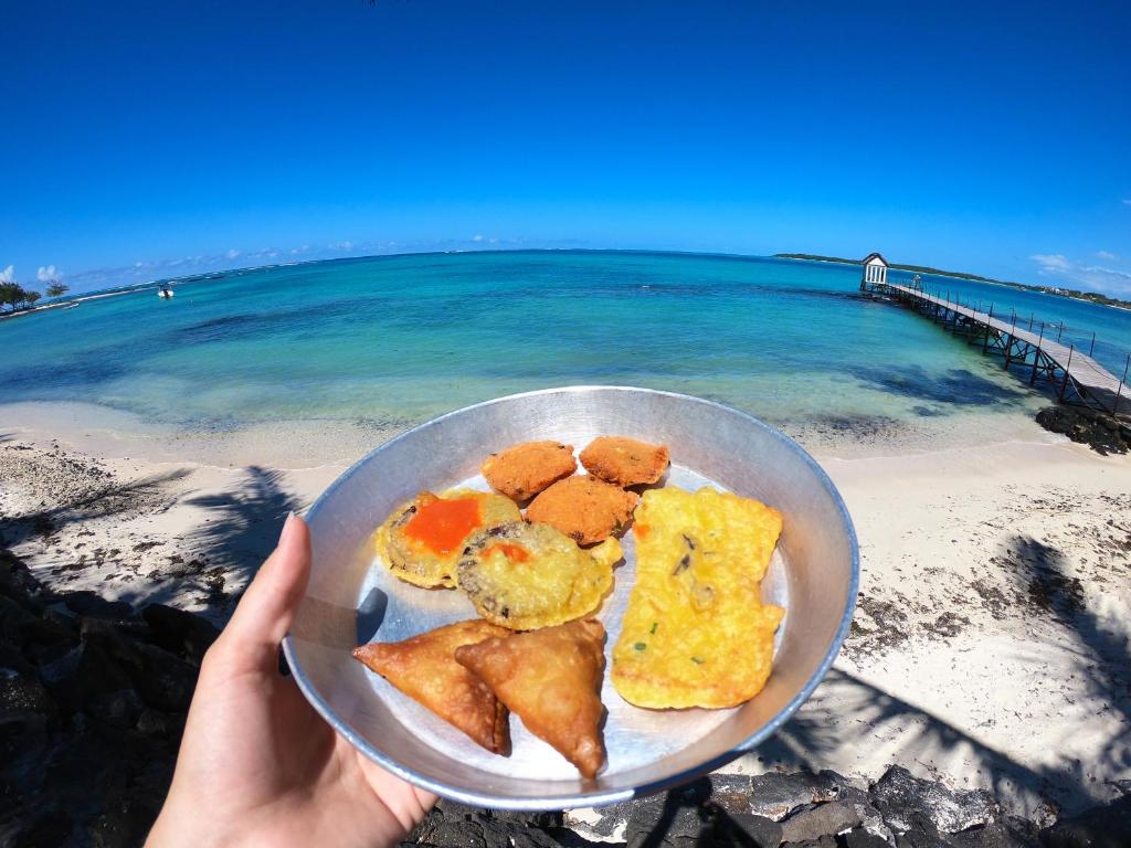 Pannā redzama plāksne ar dažādiem uzkodām, kas tiek turēta rokā, fona ir skaista pludmales ainava ar skaidru zilu ūdeni un koka tiltu.