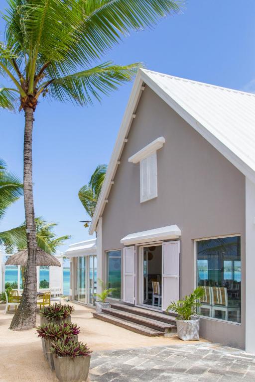 Mūsdienīga pludmales māja ar gaišu fasādi un lieliem logiem, kas piedāvā skatu uz jūru. Apkārt aug palmām un zied pētersīļi, radot relaksējošu atmosfēru.