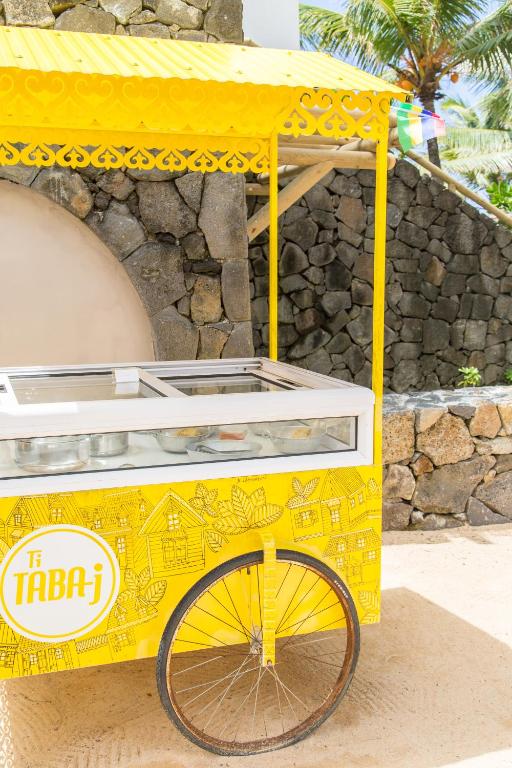 Dzeltena saldējuma rati, kas izgatavoti ar rotājumiem, atrodas pludmalē ar akmens sieniņu fonā. Rati ir aprīkoti ar stikla vitrīnu un izceļas ar krāsainu dizainu.