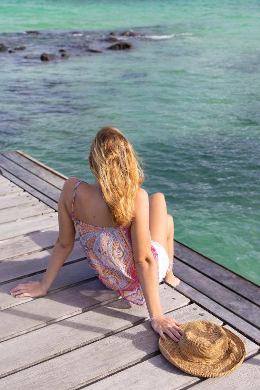 Sieviete ar gariem matiem sēž uz koka tilta, skatoties uz zaļo jūru, blakus viņai atrodas salmu cepure.