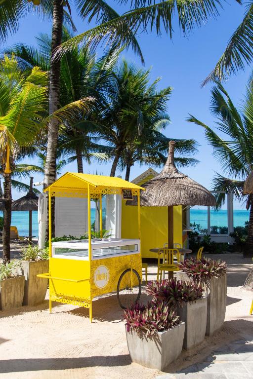 Kleita ar krāsainu dzeltenu tirdziņa stendu pludmalē, ko ieskauj palmas un ziedi. Skaista tropu vide ar zilo jūru fona.