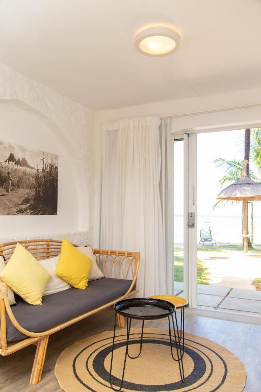 Mūsdienīgs viesistabas interjers ar rattan dīvānu un spilveniem, kas izceļas ar dzelteno krāsu. Skats uz pludmali, kur redzamas palmas un nojume.