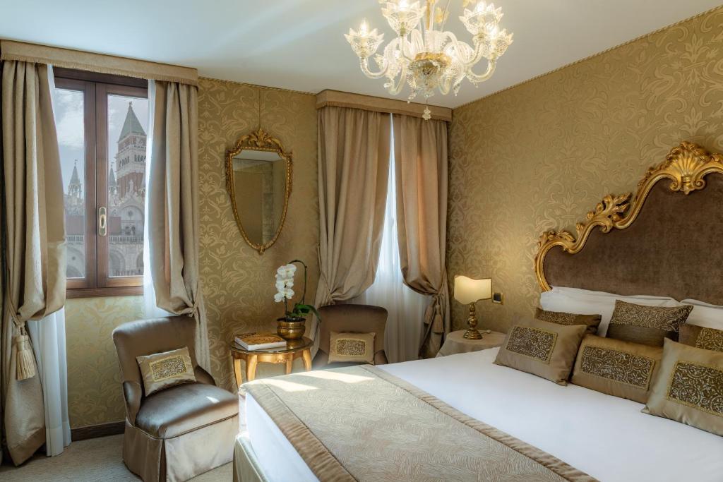 Elegants viesnīcas numurs ar greznu interjeru, zeltītiem akcentiem un panorāmas logu, caur kuru redzams pilsētas skats. Gultā ir dekoratīvas spilvendrānas, un telpā ir iekārtotas komfortablas mēbeles.