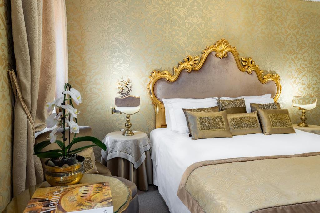 Luksusa guļamistaba ar greznu zeltītu gultas galvgalu, dekoratīviem spilveniem un elegantu interjeru. Telpā ir skaista orhideja un mājīgs apgaismojums.