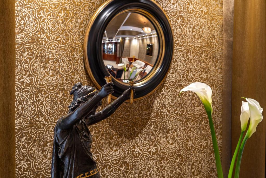 Mūsdienīgs interjera elements ar melnu spoguli un bronzas statuju, kas attēlo figūru ar loku. Fons ir izrotāts ar zeltainu rakstu, un priekšplānā redzamas baltas ziedu kāti.