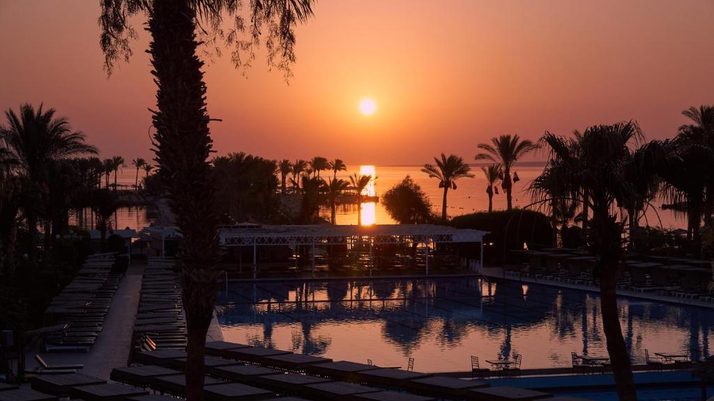 Saulriets virs baseina, apņemts ar palmām un mierīgu ūdeni, rada relaksējošu atmosfēru tropiskā kūrortā.