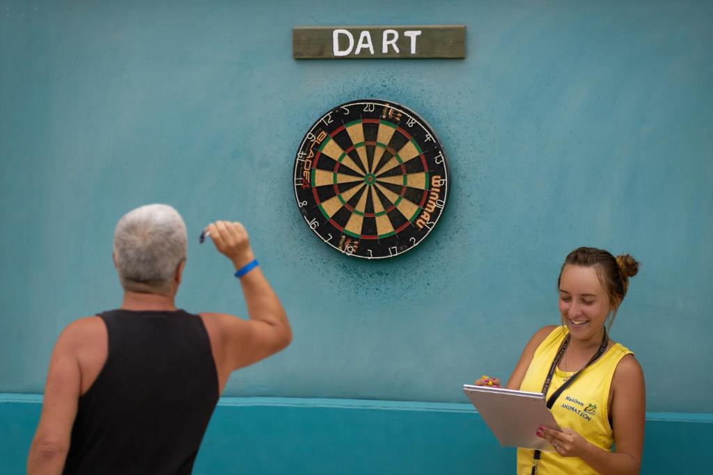 Darts spēles brīdis, kur vīrietis gatavojas mest, bet sieviete fiksē rezultātus. Sienā redzama mērķa tablo ar uzrakstu "DART".