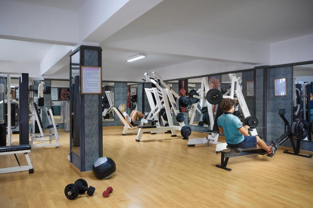 Sporta zāle ar dažādām trenažieru iekārtām un cilvēkiem, kas nodarbojas ar fiziskajām aktivitātēm. Telpa ir gaiša un plaša, ar koka grīdu un spoguļiem uz sienām.