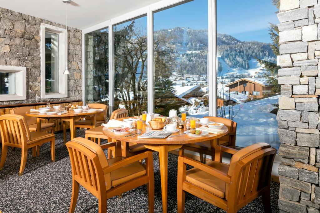 Skaista ēdamzāle ar koka mēbelēm, kurā ir galds, kas klāts ar brokastu ēdieniem un dzērieniem, un caur lieliem logiem paveras skats uz sniegotajiem kalniem un mājām.