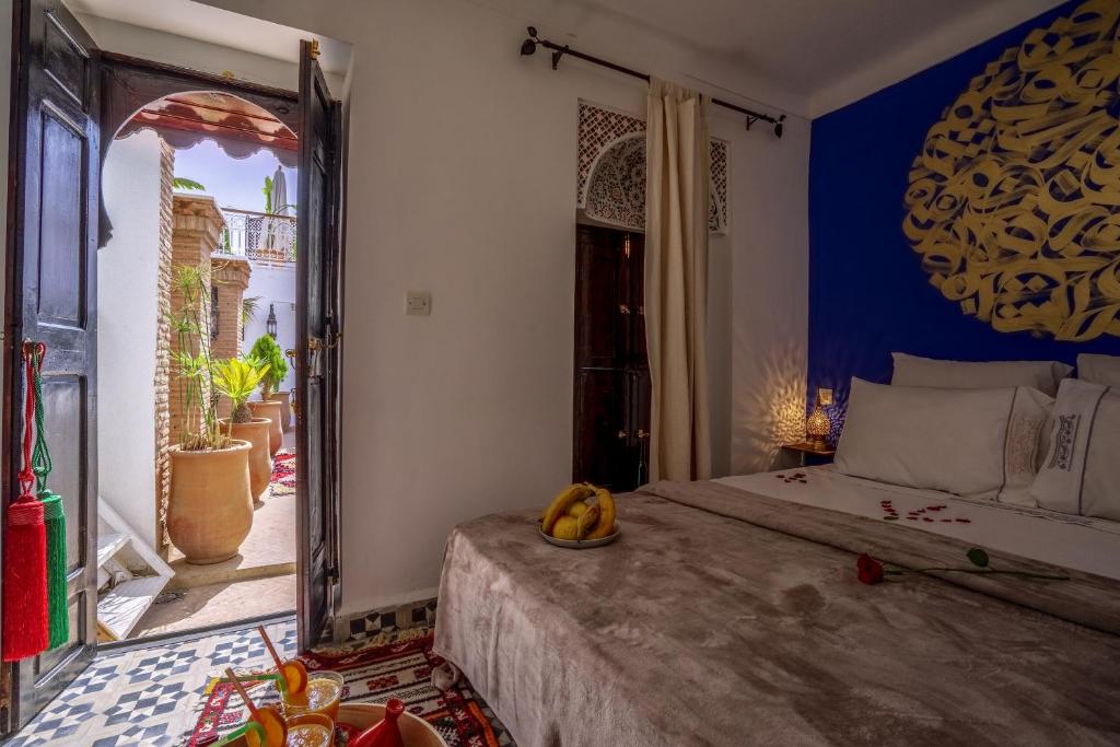 Mūsdienīgs viesnīcu numurs ar zilu sienu un greznu dekoru, kur logs ved uz zaļu terasi ar puķēm. Gultā ir spilveni un ziedi, radot romantisku atmosfēru.