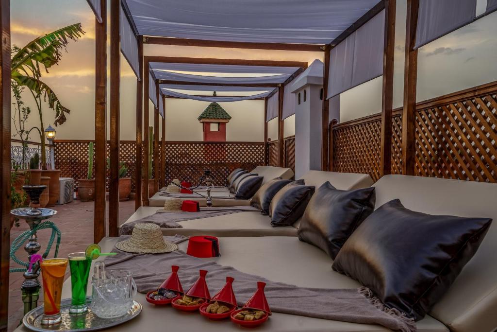 Relaksējoša terase ar komfortablām gultām, uz kurām ir spilveni, un dažādiem dzērieniem un uzkodām uz galda. Fons ar saulrietu un dekoratīvām augu kompozīcijām rada mierīgu atmosfēru.