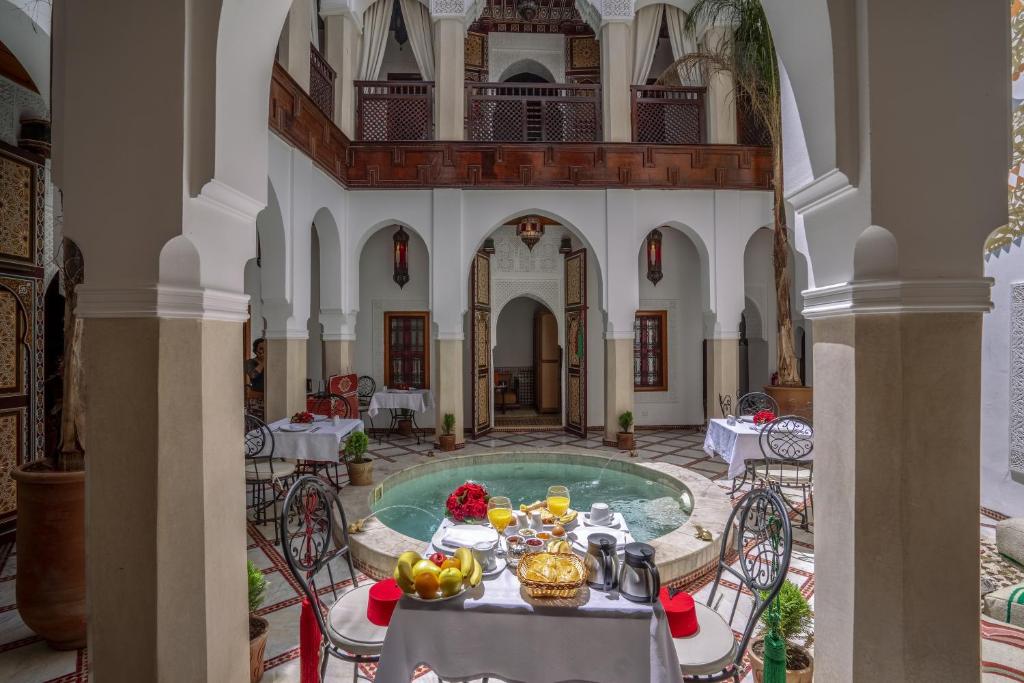 Elegants iekšējs pagalms ar tradicionālu marokāņu dizainu, kurā ir rakstainā flīze un ūdens baseins. Uz galda ir izliktas brokastu ēdienu plāksnes, augļi un dzērieni, radot viesmīlīgu atmosfēru.