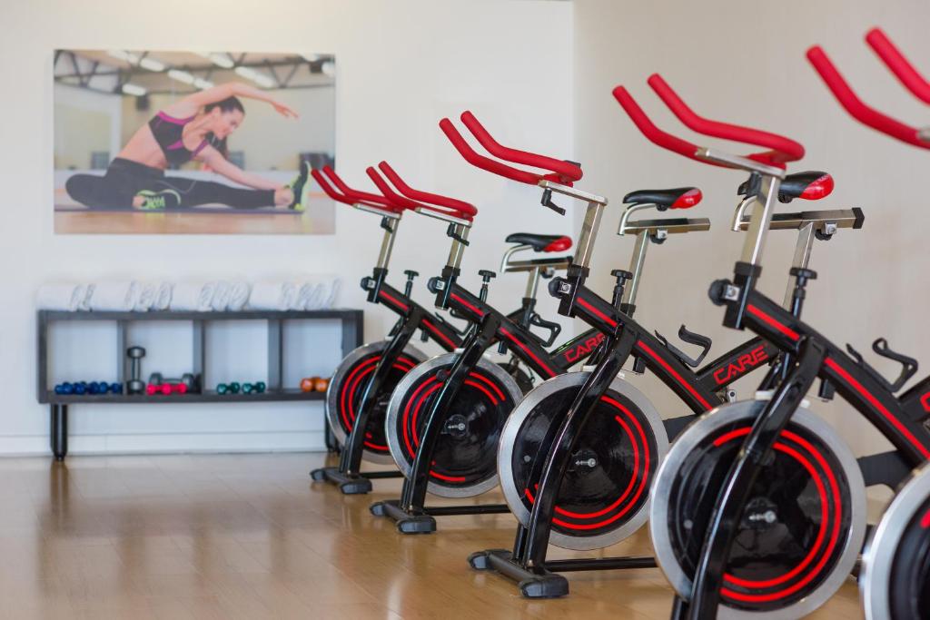 Sporta zāles interjers ar velosipēdu trenažieriem un vingrošanas aprīkojumu, kas aicina uz aktīvu dzīvesveidu. Fons rotā attēls ar sportistiem, kas izstaro enerģiju un veselību.