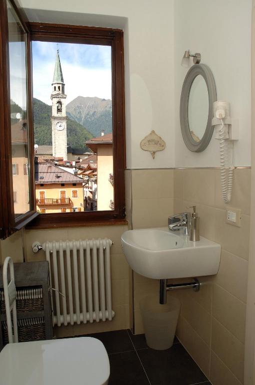 Vannas istaba ar logu, kas sniedz skatu uz kalnu pilsētu un zvanu torni, nodrošinot mierīgu atmosfēru. Redzams moderns izlietnes dizains un apkures radiators.