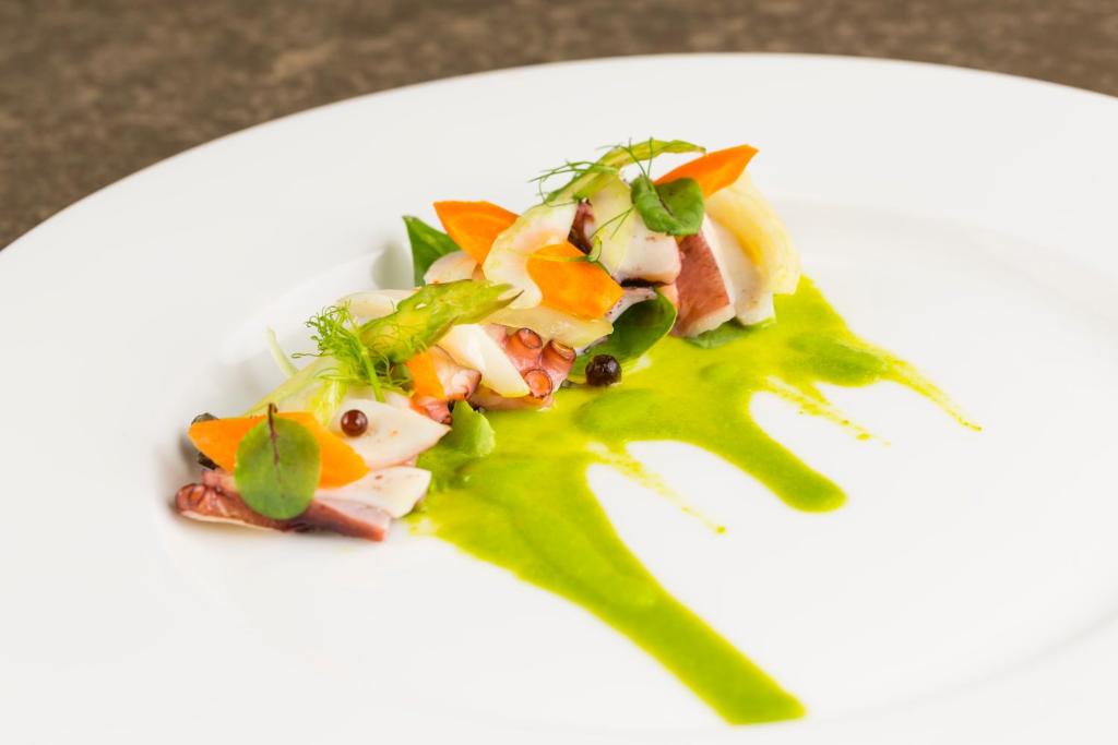 Gourmet jūras veltes uz balta šķīvja ar dārzeņu dekorējumu un zaļo mērci. Šis elegantais ēdiens izceļas ar krāsainām sastāvdaļām un radošu pasniegšanas veidu.