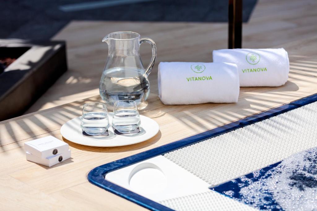 Stilīgs relaksācijas stūrītis ar ūdens karafēm, divām glāzēm un VITANOVA dvieļiem, kas novietoti uz koka virsmas blakus baseinam.