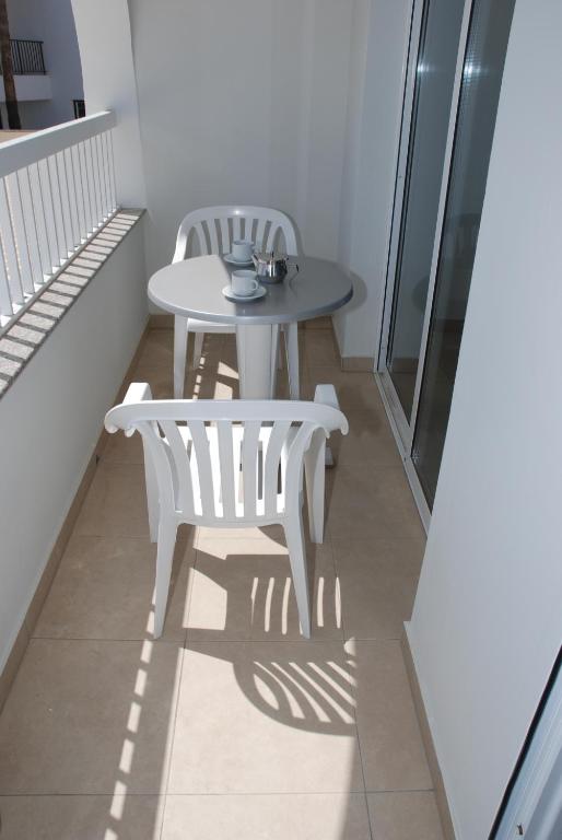 Balcony ar apaļu galdu un divām baltām plastmasas krēsliem, uz galda ir kafijas tasīte un cukura trauks. Gaisma veido ēnas uz flīzēm, radot mierīgu atmosfēru.