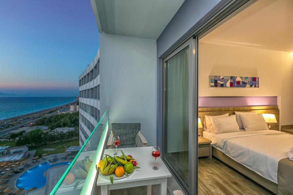 Balcony view from a hotel room featuring a table with fresh fruit and drinks, overlooking the sea and a pool area at sunset.