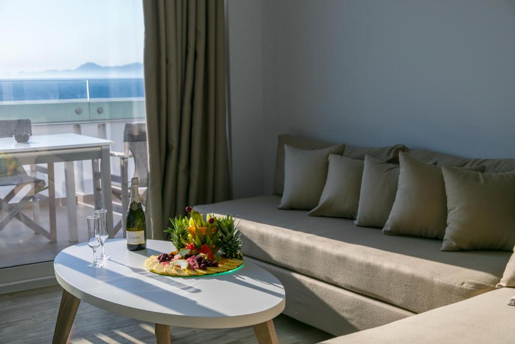 Mūsdienīga viesnīcas istaba ar skatu uz jūru, kurā redzama dīvāna zona, galda ar šampanieti un augļu šķīvi. Gaisīga atmosfēra un komforts, kas aicina atpūsties.