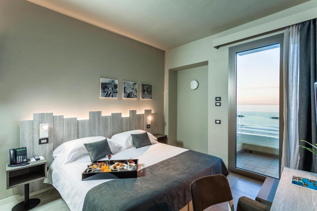 Mūsdienīgs viesnīcas numurs ar skatu uz jūru, ērts gultas komplekts un brokastu šķīvis uz galda. Gaisīga atmosfēra ar lieliem logiem, kas ļauj ieplūst dabiskajai gaismai.