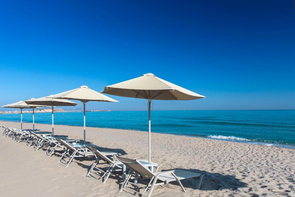 Pludmales skats ar saulessargiem un sauļošanās krēslām, kas izvietoti smilšainā piekrastē, ar mierīgu jūru un skaidru zilo debesu fonu.