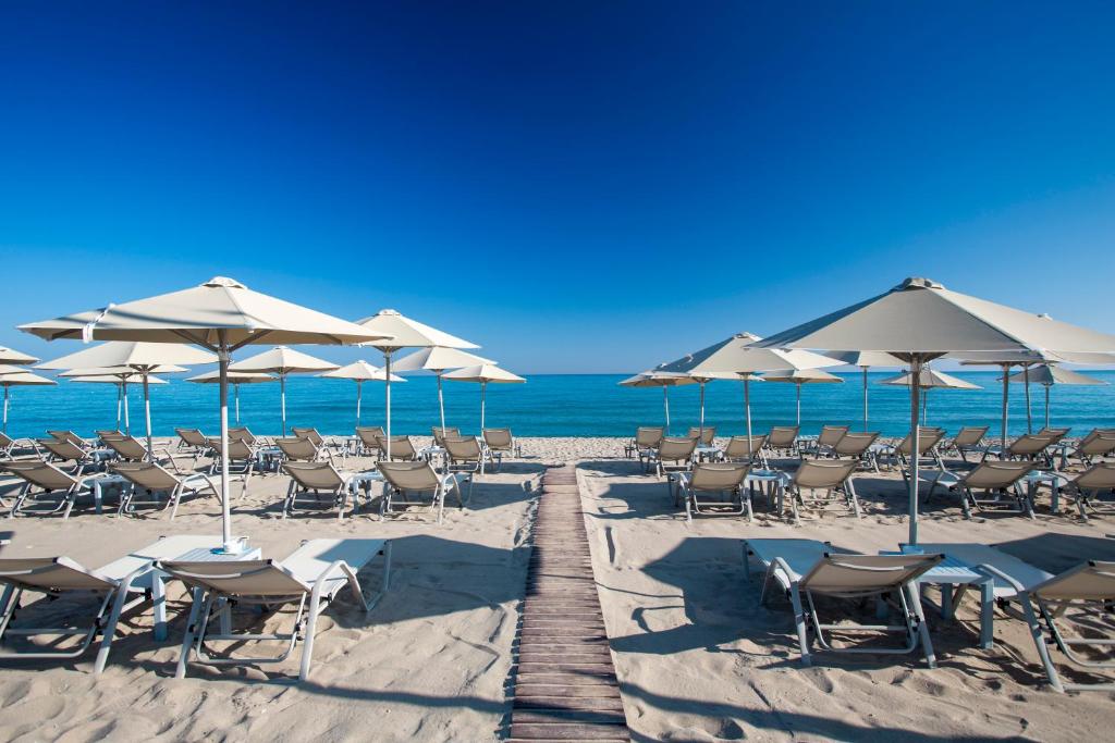 Pludmales skats ar saulessargiem un atpūtas krēsliem, kas novietoti smiltīs pie mierīga ūdens, zem skaidra zilā debesīm.