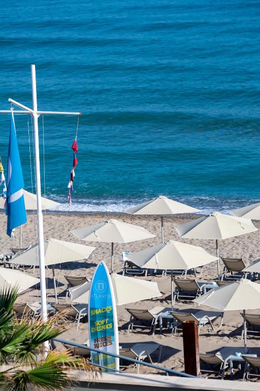 Pludmales skats ar saulessargiem un krēsliem, blakus zilo ūdeņu krastam, kur redzama zīme ar uzrakstu "Uzkodas, dzērieni, bezmaksas sulas".