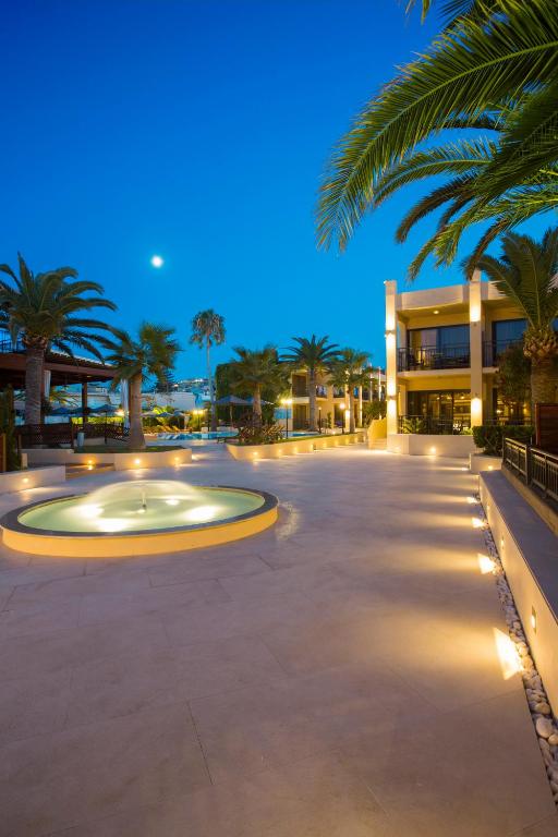 Mūsdienīgs kūrorts ar plašu terasi un baseinu, apgaismotu ar siltu gaismu, tropisko augu un palmu fonā, pie vakara debesīm.