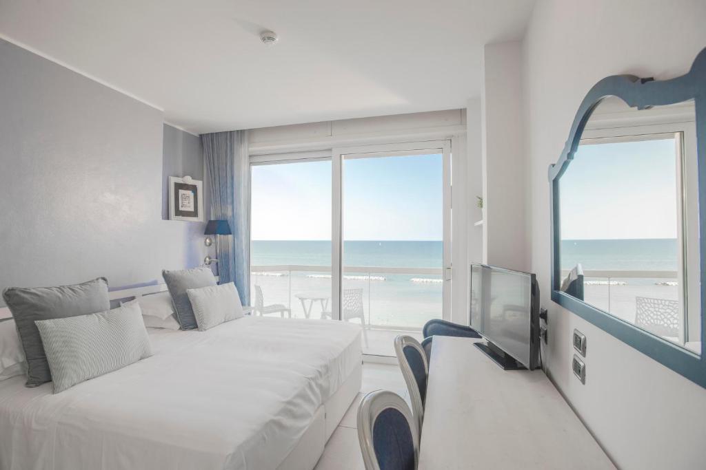 Gaiša viesnīcas istaba ar divām gultām un skatu uz jūru, kurā ir televizors un balta rakstāmgalda zona. Logi ir atvērti, ļaujot ieplūst dabiskajai gaismai.