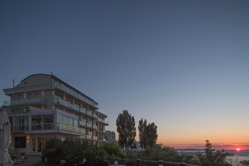 Mūsdienīga viesnīca pie jūras, kurā redzams saulriets un skaidrs debesis. Apkārt viesnīcai ir zaļš apstādījums un koku rinda.