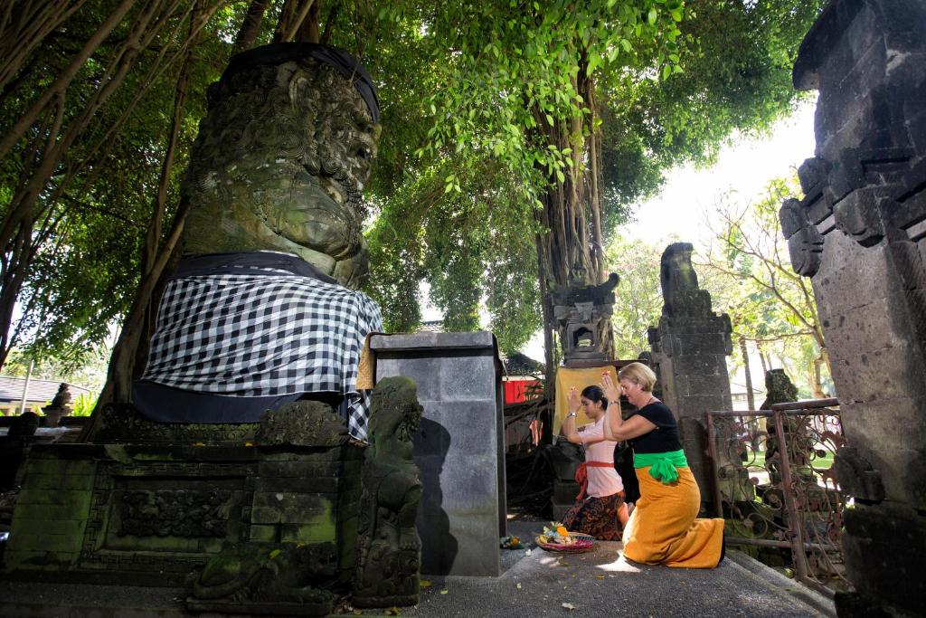 Divas sievietes lūdz, stāvot pie akmens statujas, kas ir apģērbta ar melnbaltu audumu, starp zaļām koku lapām un akmens struktūrām.