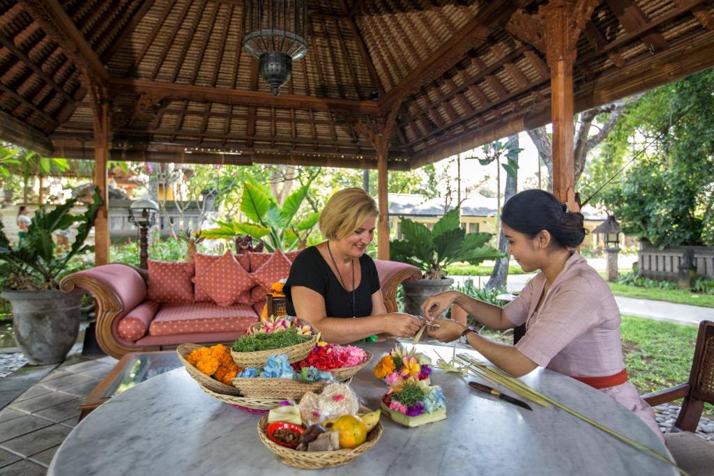 Divas sievietes, sēžot zem tradicionālas jumta konstrukcijas, izgatavo ziedu rotājumus, izmantojot krāsainus ziedus un augļus, kam blakus ir greza dīvāna zona un sulīgi zaļumi.