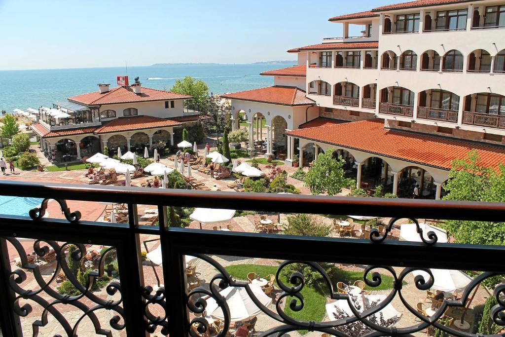 Skats no balkona uz viesnīcas dārzu un jūru, kur redzami sauļošanās krēsli un baseins. Viesnīca ar sarkaniem jumtiem un zaļām stādījumām piedāvā relaksējošu atmosfēru.