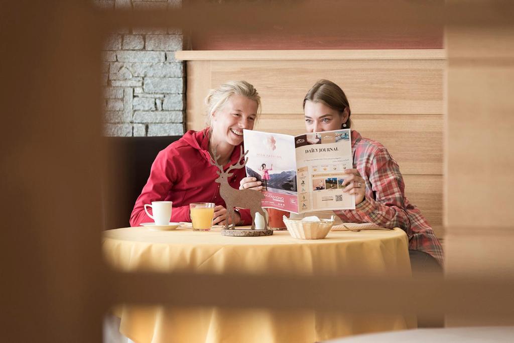 Divas sievietes sēž pie galda, skatoties ēdienkarti un smejoties, kamēr uz galda ir dzērieni un maizes groziņš. Gaisotne ir draudzīga un relaksēta.
