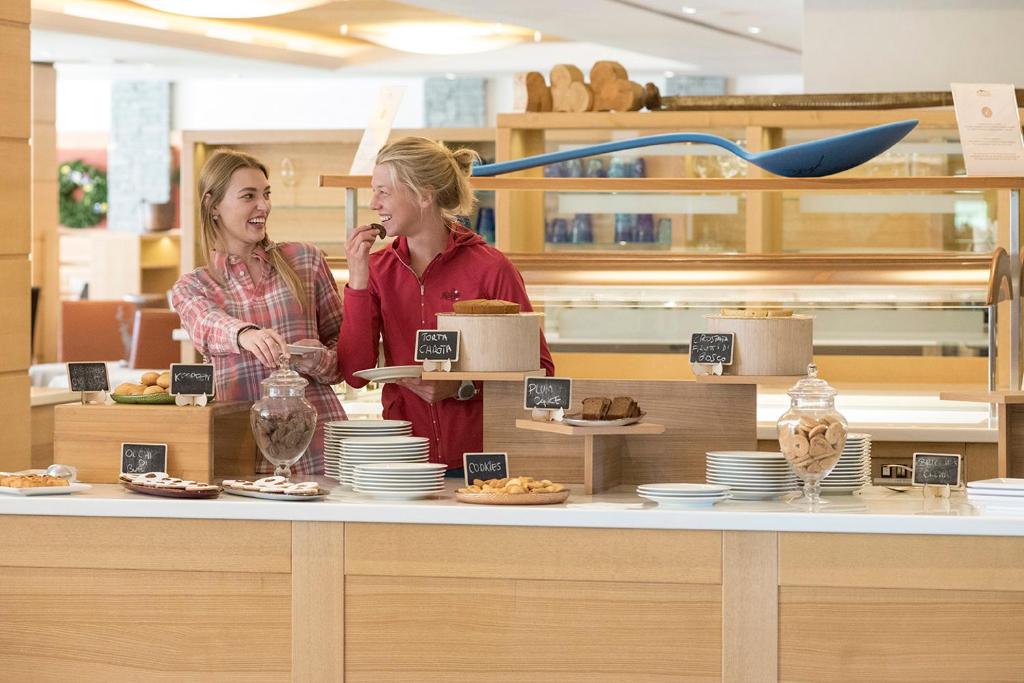 Divas sievietes priecīgi sarunājas un gardēžojas pie bufetes, kurā ir dažādi cepumi un saldumi. Gaisotne ir silta un draudzīga, ideāla brīvdienu baudīšanai.