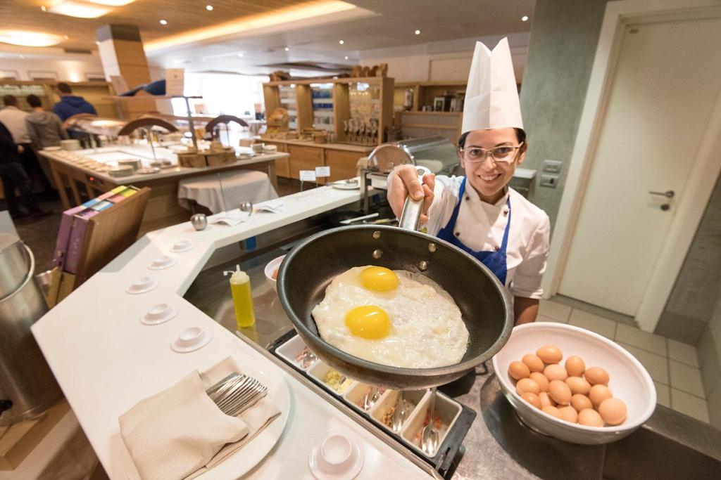 Šefpavārs, kurš pasniedz divas ceptas olas pannā, stāv pie bufetes, kurā redzami daudzi olu trauki un citas ēdienu sagatavošanas zonas.